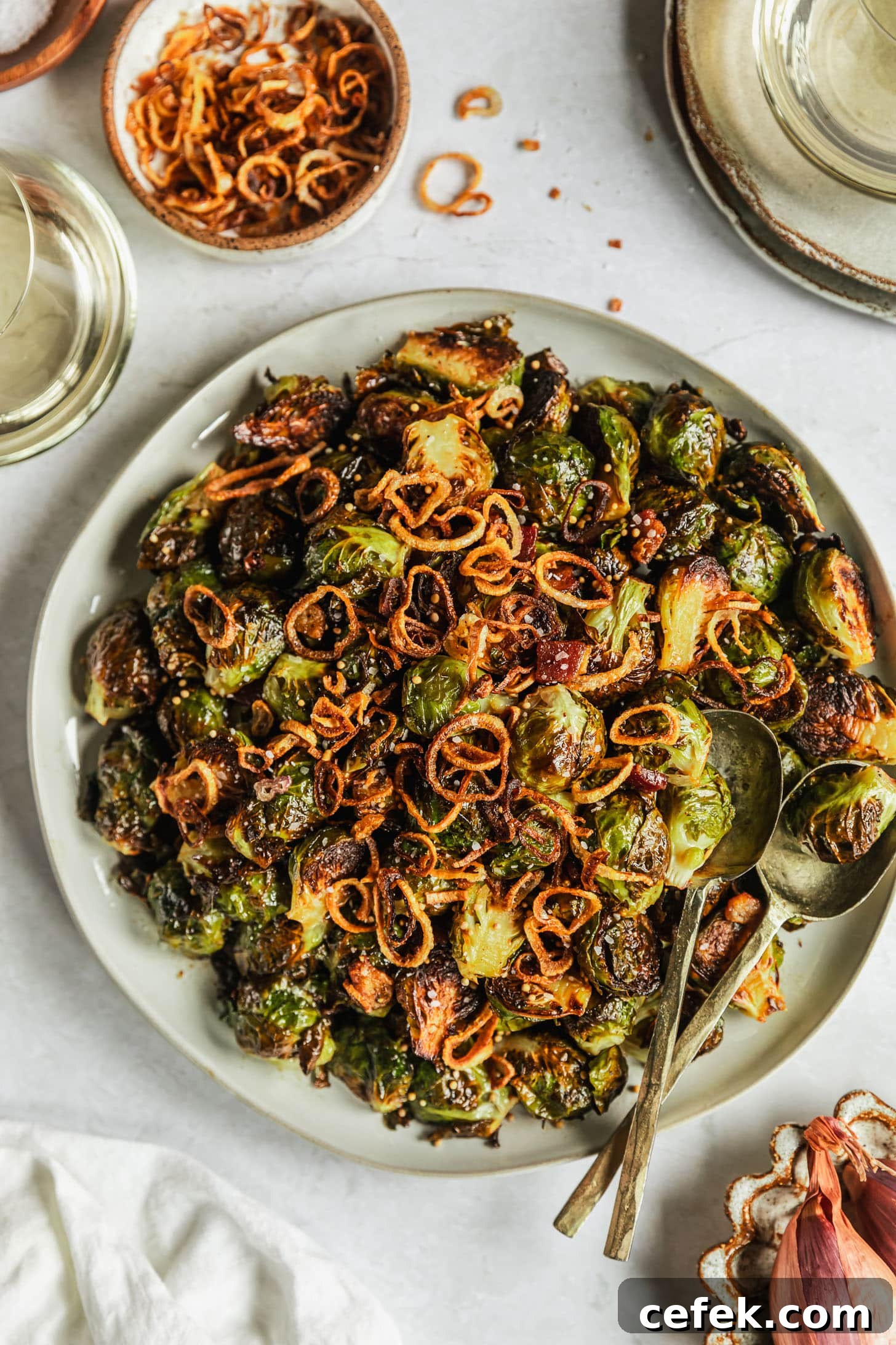 A white plate of caramelized Brussels sprouts with bacon on a white marble counter next to two glasses of white wine, white bowl of fried shallots, and white linen.