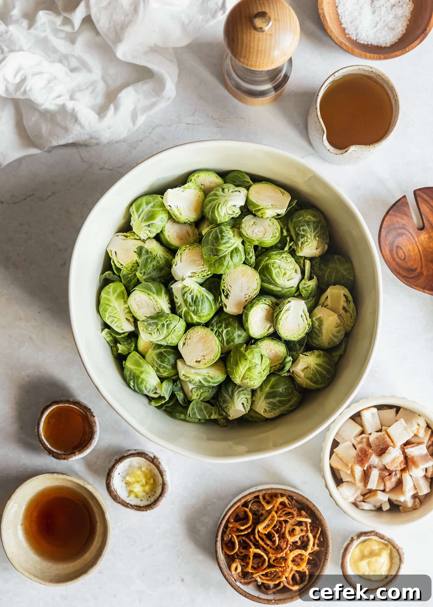 White bowls of Brussels sprouts, raw bacon, fried shallots, Dijon, honey garlic, vinegar, and oil on a white marble counter next to a white linen and wood spoon.