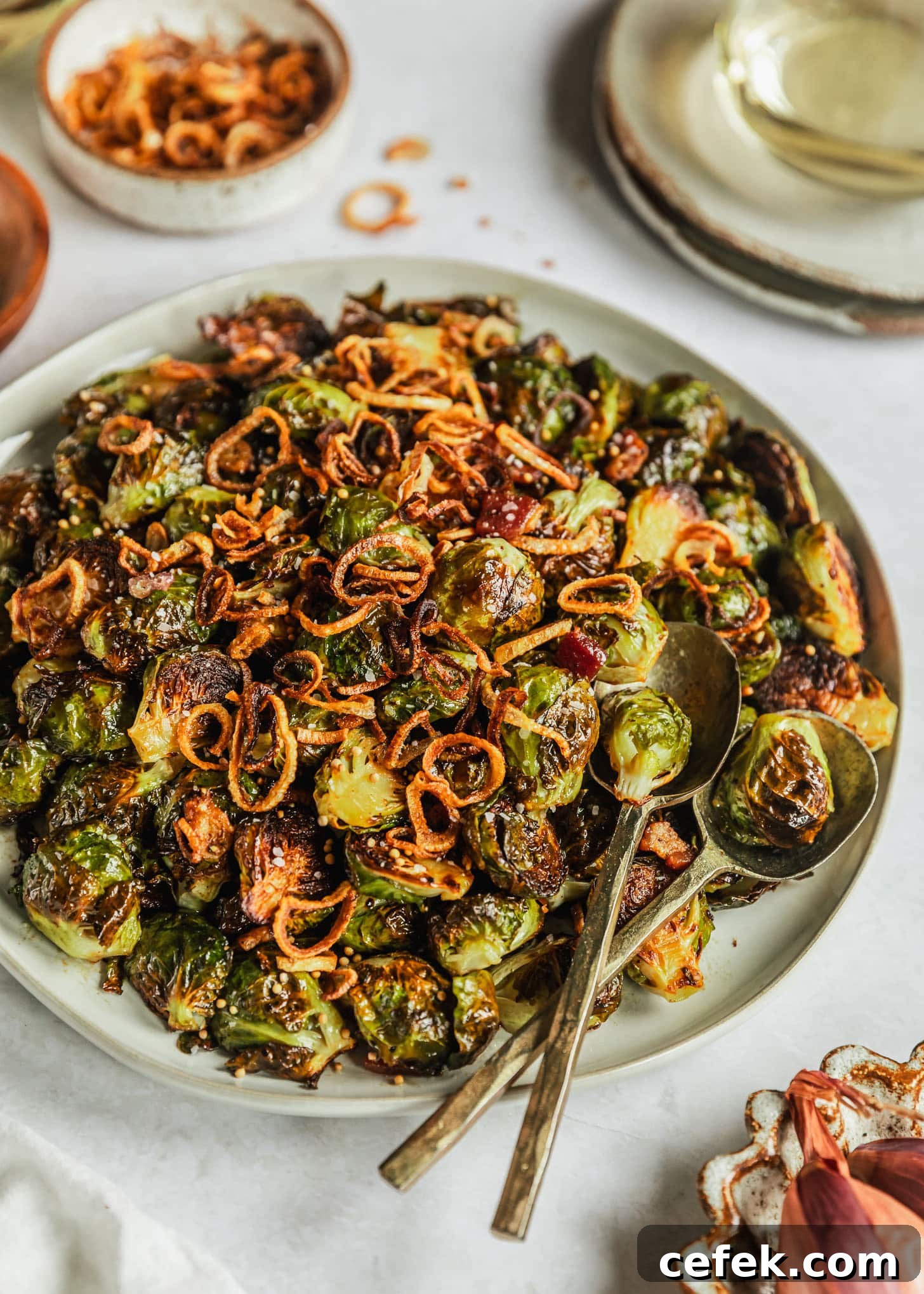 A white platter of caramelized Brussels sprouts with bacon on a white marble counter next to a white bowl of fried shallots, grey plates, and a white bowl of shallots.
