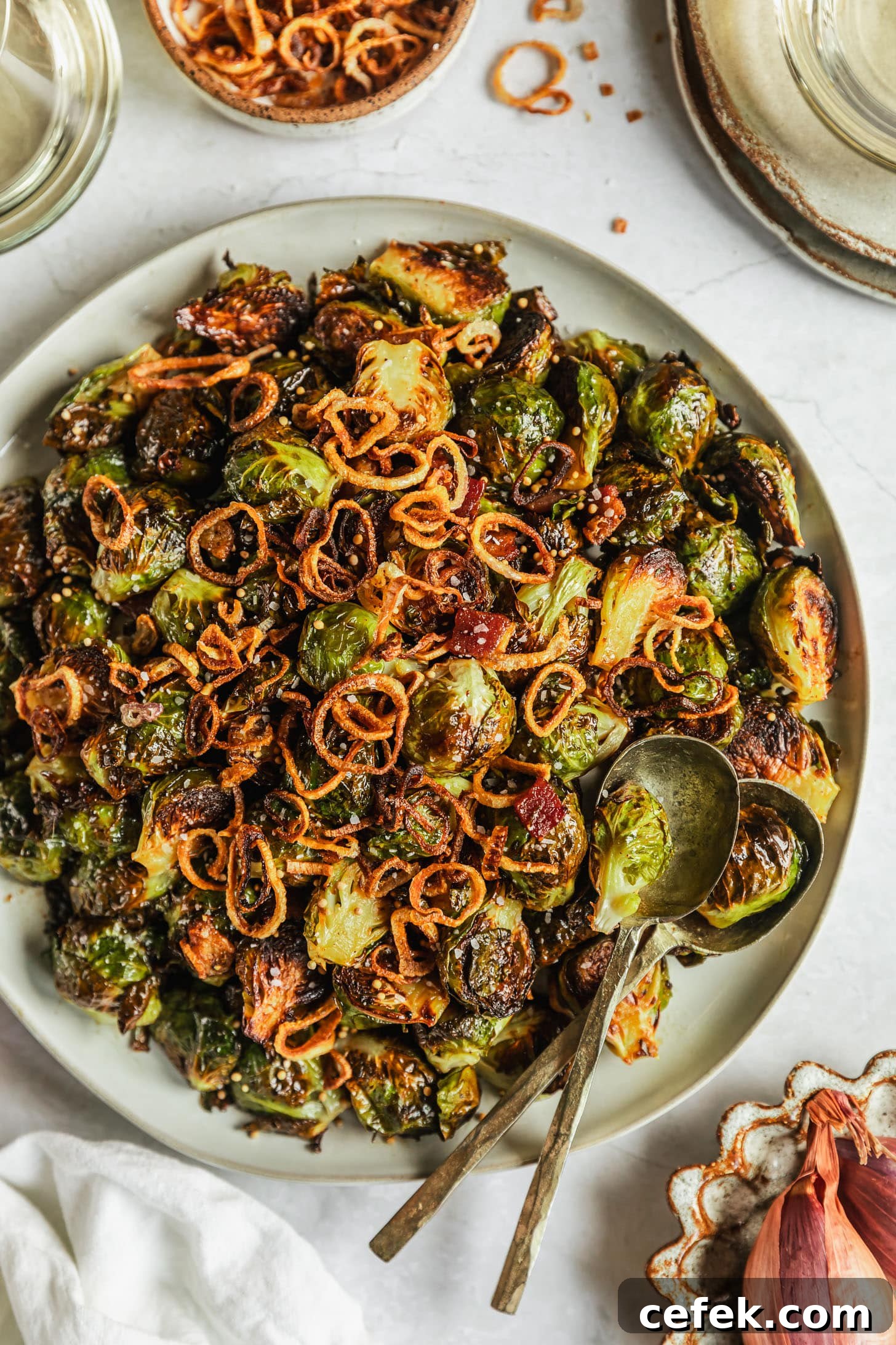 A white platter of crispy caramelized Brussels sprouts with bacon on a marble counter next to grey plates, a white linen, two glasses of white wine, and a white bowl of fried shallots.