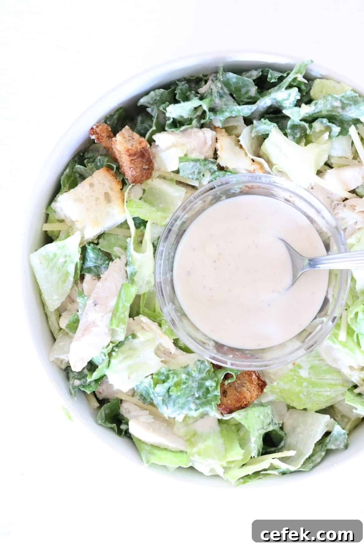 A small glass bowl with Caesar dressing with a spoon in the middle of a bowl of Caesar salad.