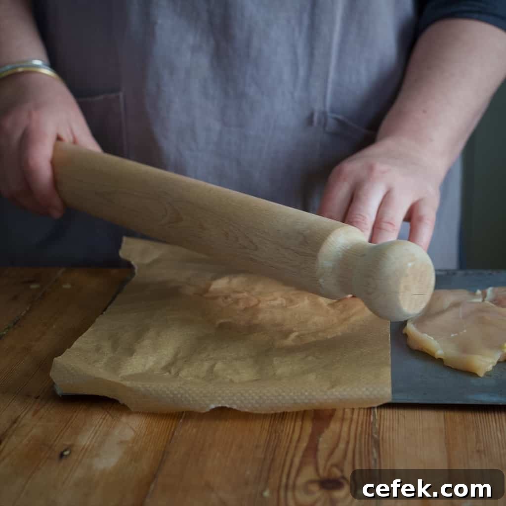 Woman in grey flattening a butterflied chicken breast on a chopping board with a wooden rolling pin