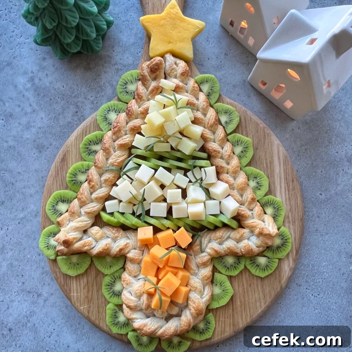 A Christmas tree-shaped pastry topped with cubes of cheese and surrounded by kiwi slices, displayed on a round wooden board.