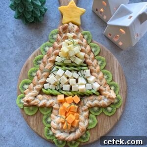 A Christmas tree-shaped pastry braid topped with cheese cubes and surrounded by kiwi slices on a wooden board, with decorative ceramic houses in the background.
