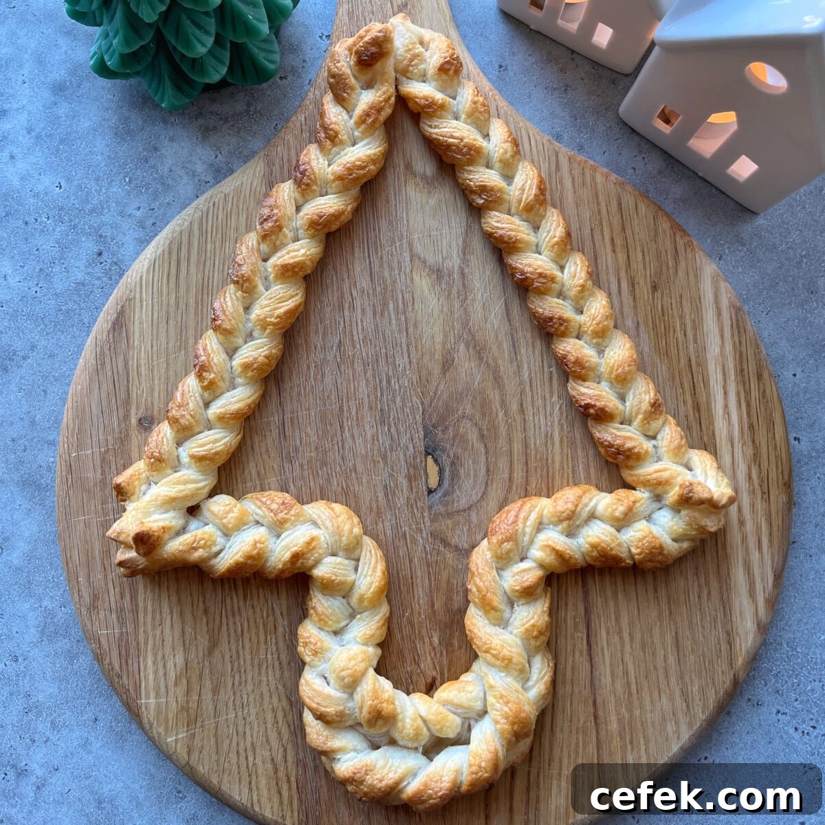 A braided pastry shaped like a Cheese Board Christmas Tree sits on a round wooden board, with decorative houses and a green tree nearby.