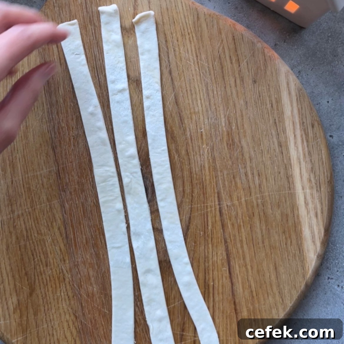 Three strips of dough are laid out side by side on a round wooden cutting board, with a hand reaching toward them from the left.
