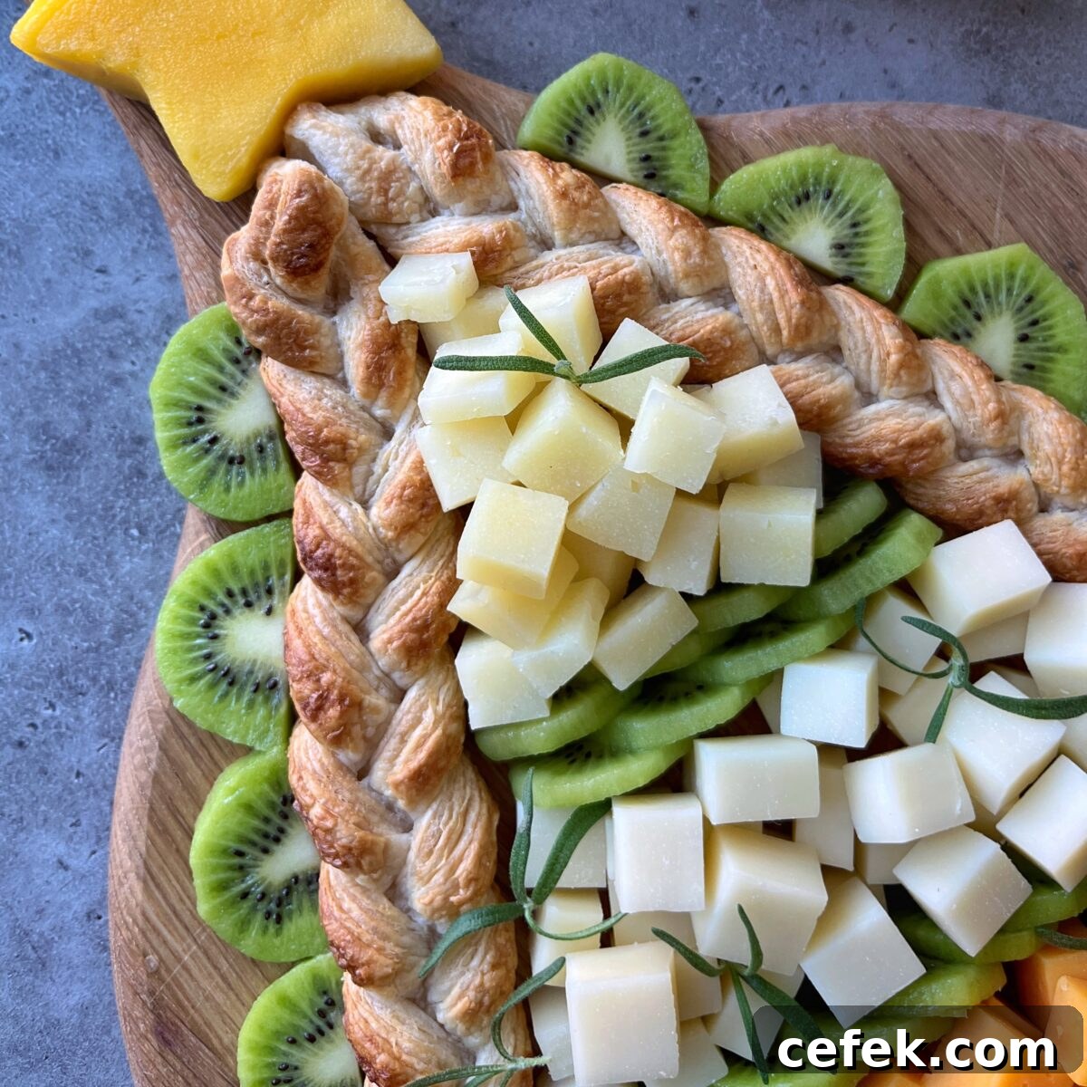 A cheese board is arranged decoratively with sliced kiwi, cubed white cheese, braided puff pastry, and a star-shaped cheese piece on top.