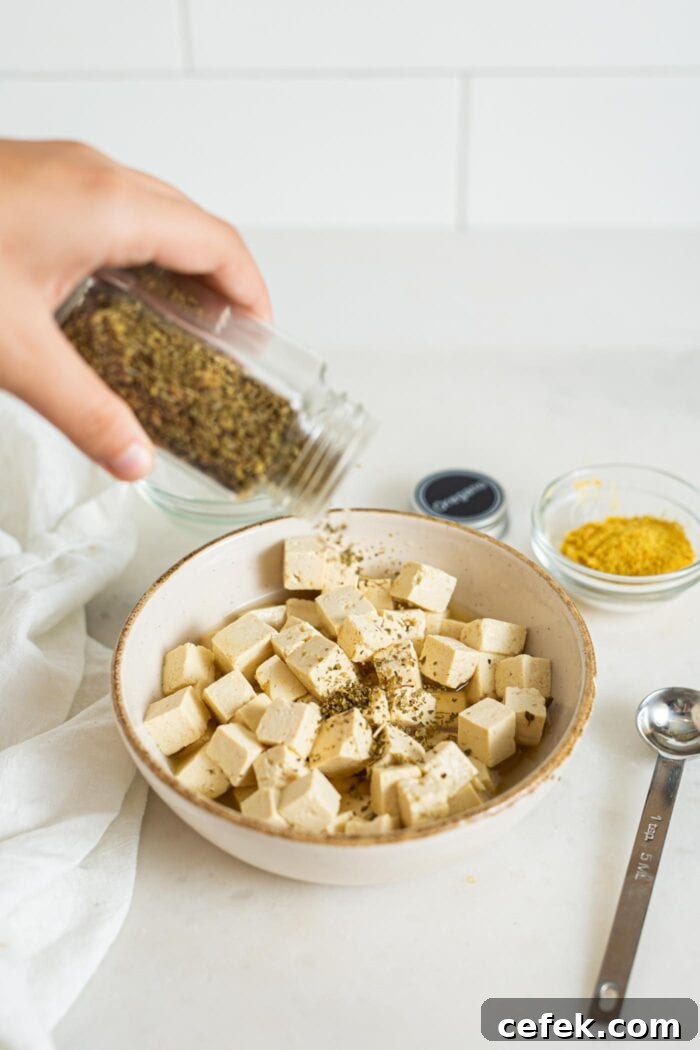 Adding dried oregano to a bowl of cubed tofu feta. Spoon and dish of nutritional yeast beside the bowl.