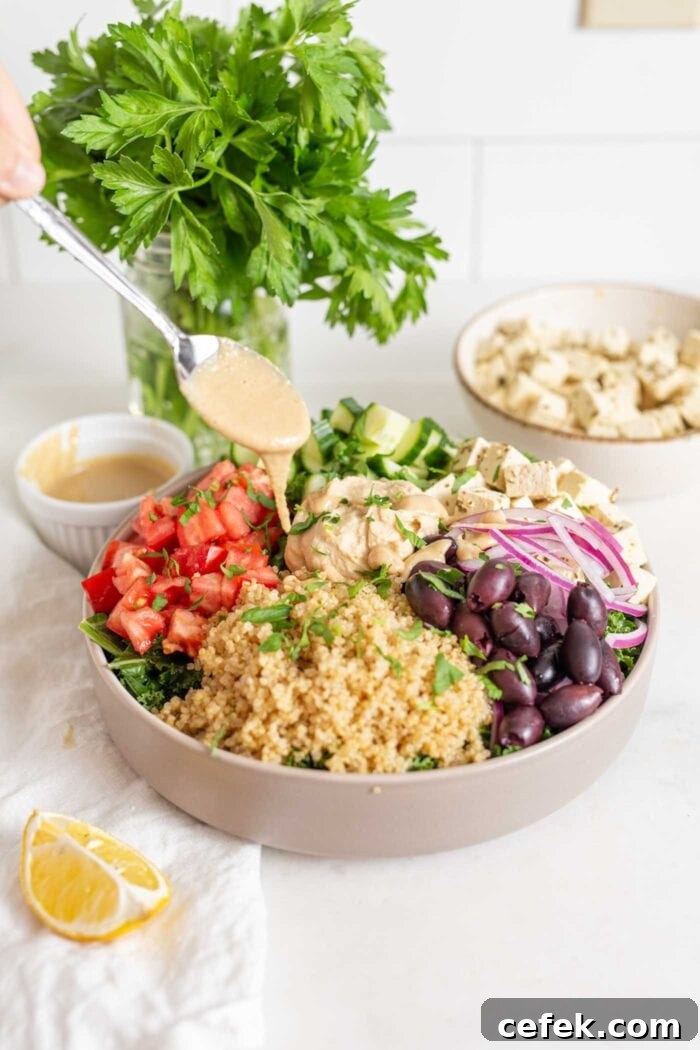 Adding a spoonful of tahini to a olorful bowl of quinoa, chopped tomato and peppers, red onion, olives and tofu.