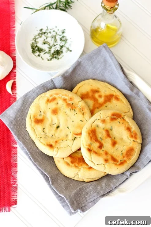 4 Rosemary and Garlic Flatbread rounds sitting on a gray napkin on a white table, olive oil, garlic and rosemary on side.