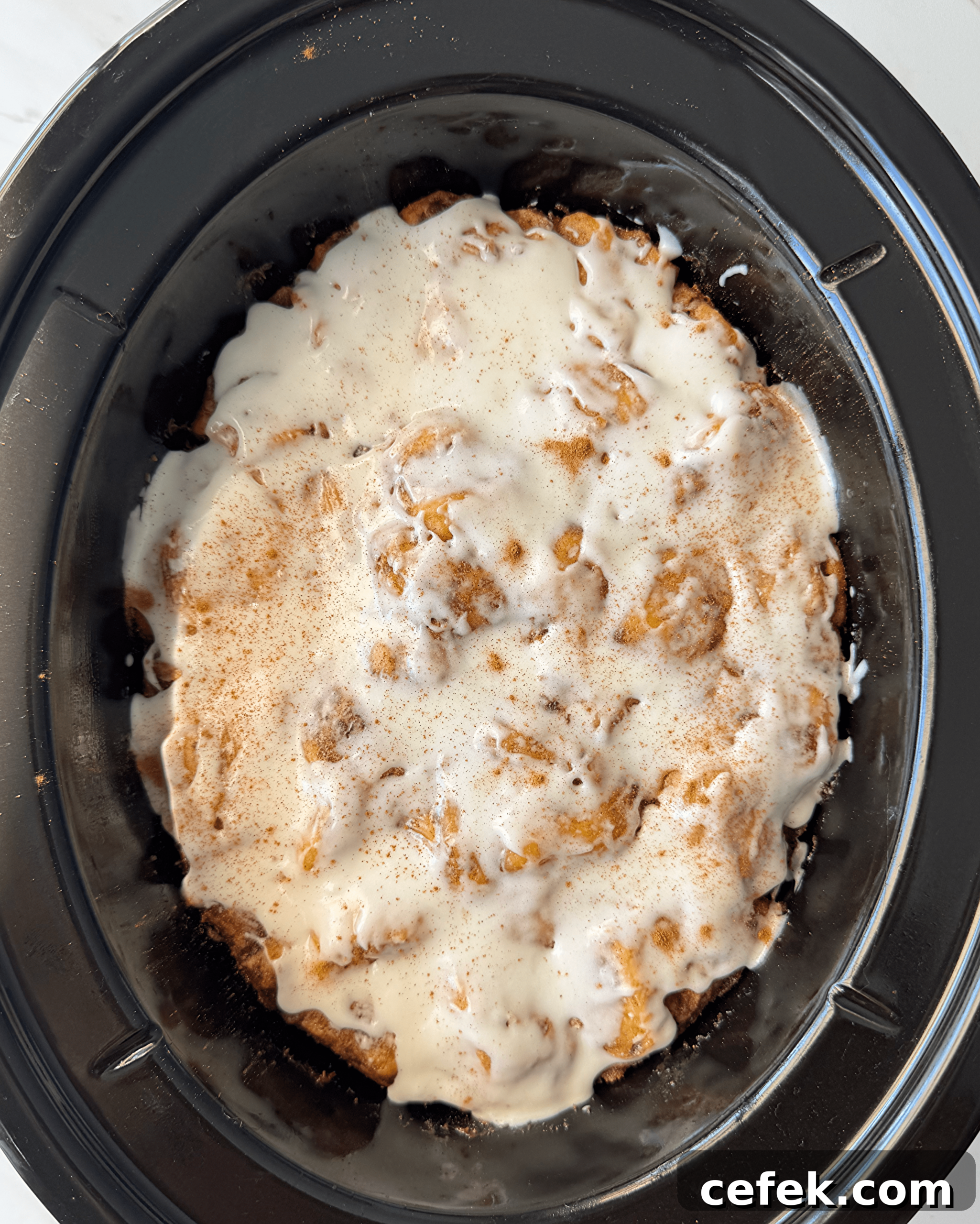 overhead shot of cooked Cinnamon Roll Casserole in a black slow cooker with icing and cinnamon sprinkled on top