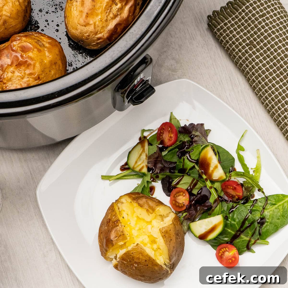 A baked potato cut open with butter in the middle served with a side salad and a slow cooker in the background with jacket potatoes in the bottom of the cooker