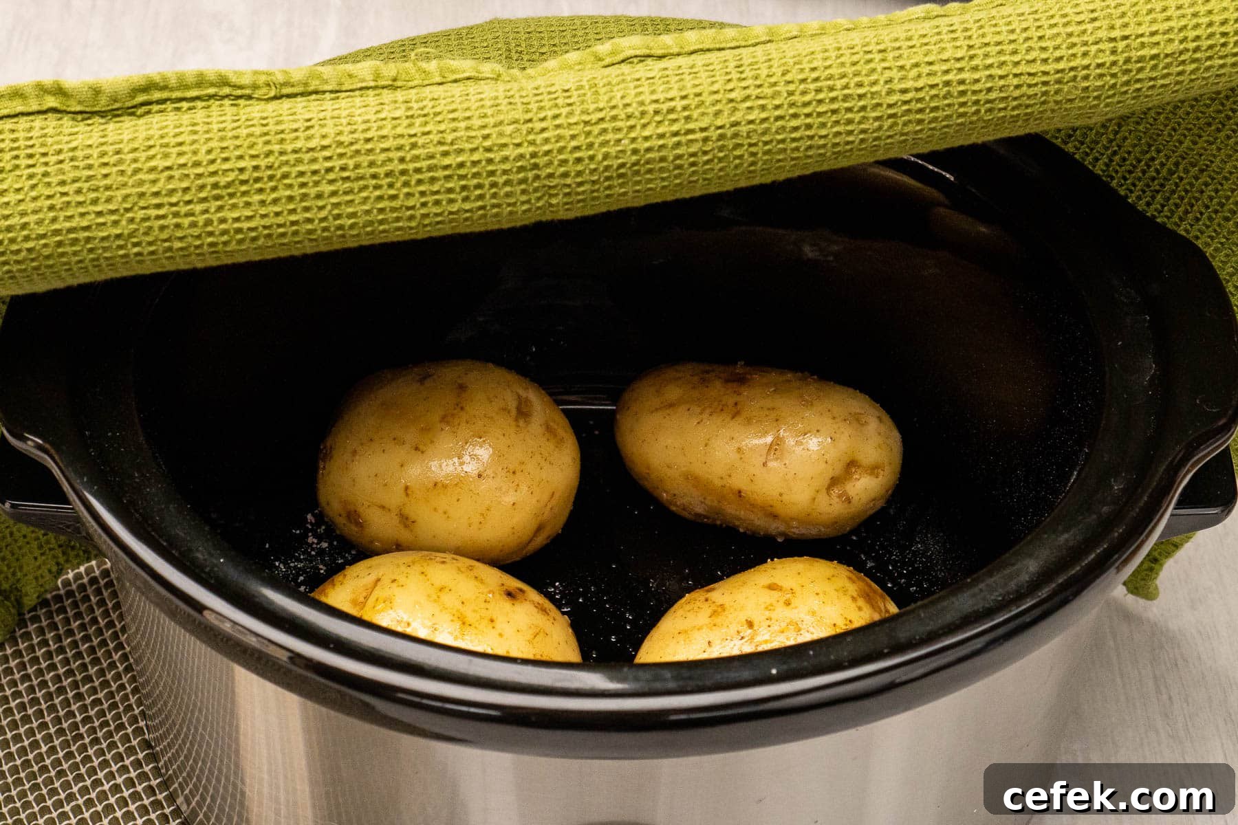 Placing a clean green tea towel over the top of a slow cooker with four potatoes in the bottom of the cooker