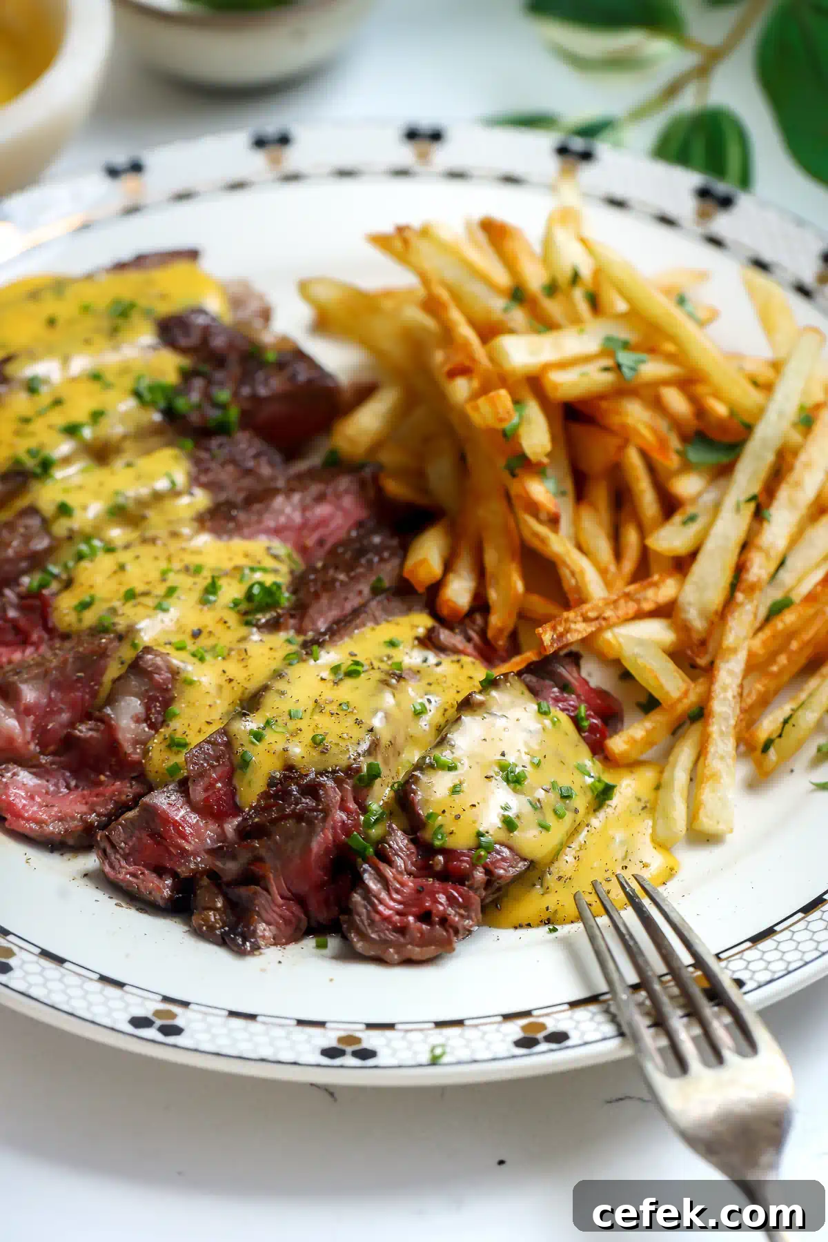 Close up of sliced steak drizzled with herb bearnaise sauce next to a pile of thin crispy frites with a silver fork in view.