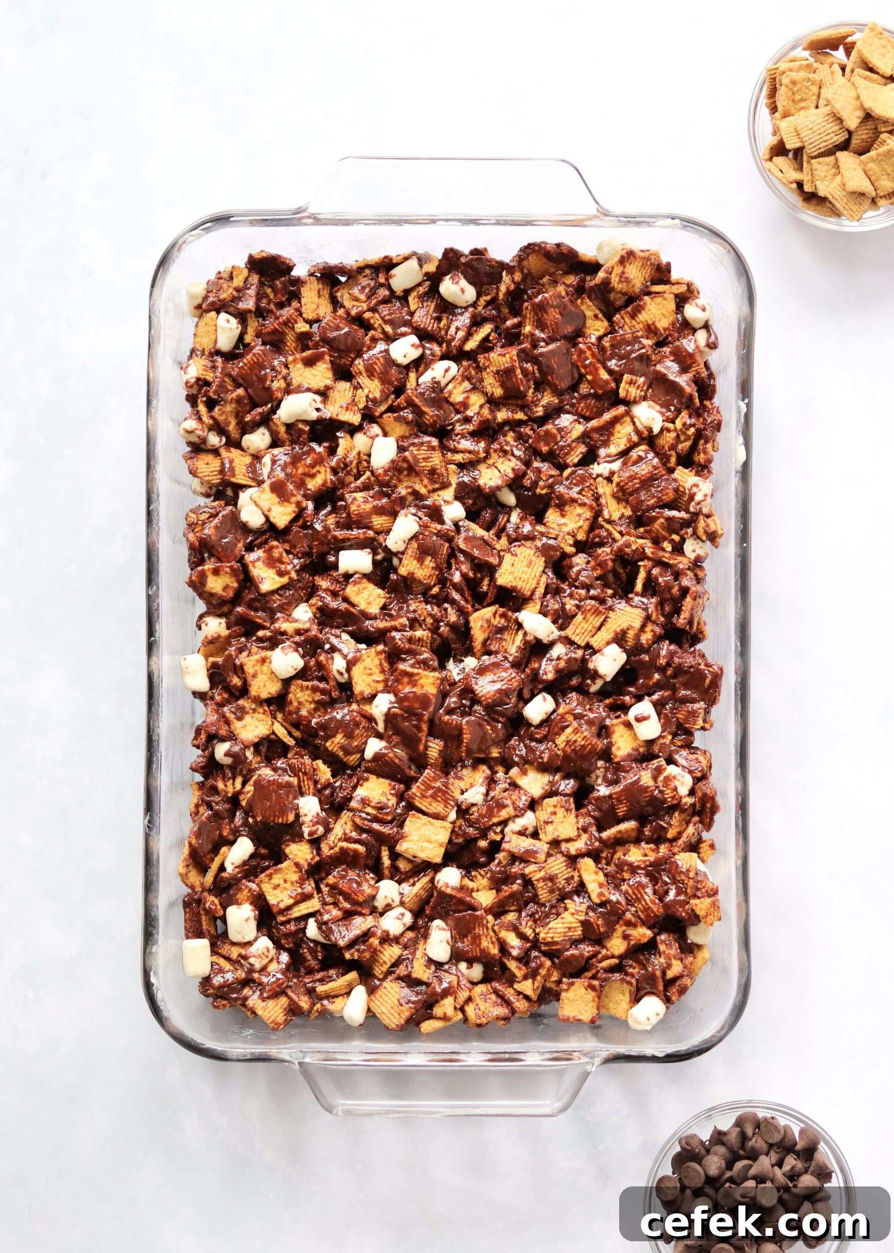 Cereal s'mores mixture pressed into a baking dish with small bowls of chocolate chips and cereal next to it.