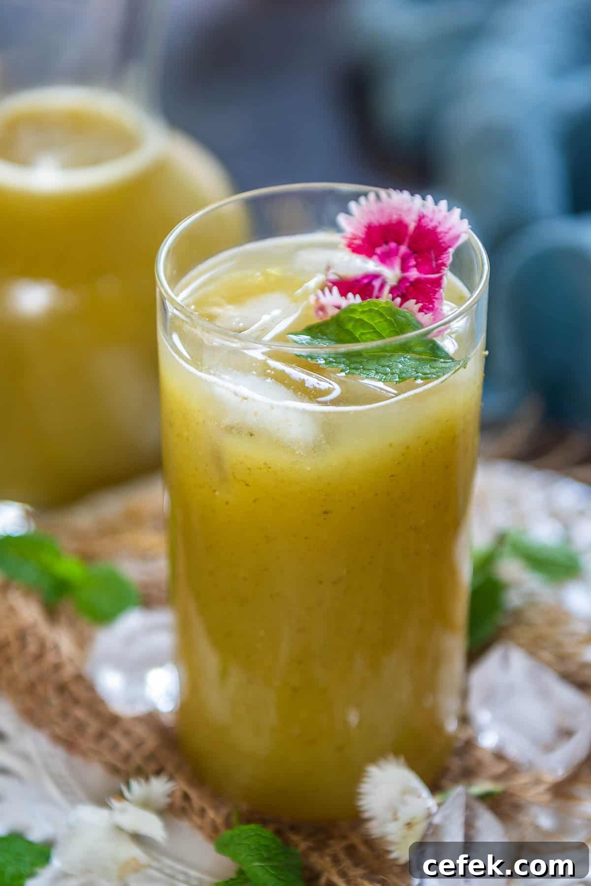 A tall glass of yellow-green Aam Panna drink with ice cubes, garnished with a sprig of mint and pink edible flowers, set on a rustic surface with scattered mint leaves and ice.