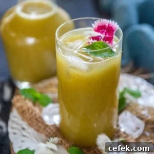 A tall glass of yellow-green Aam Panna drink with ice cubes, garnished with a sprig of mint and pink edible flowers, set on a rustic surface with scattered mint leaves and ice.