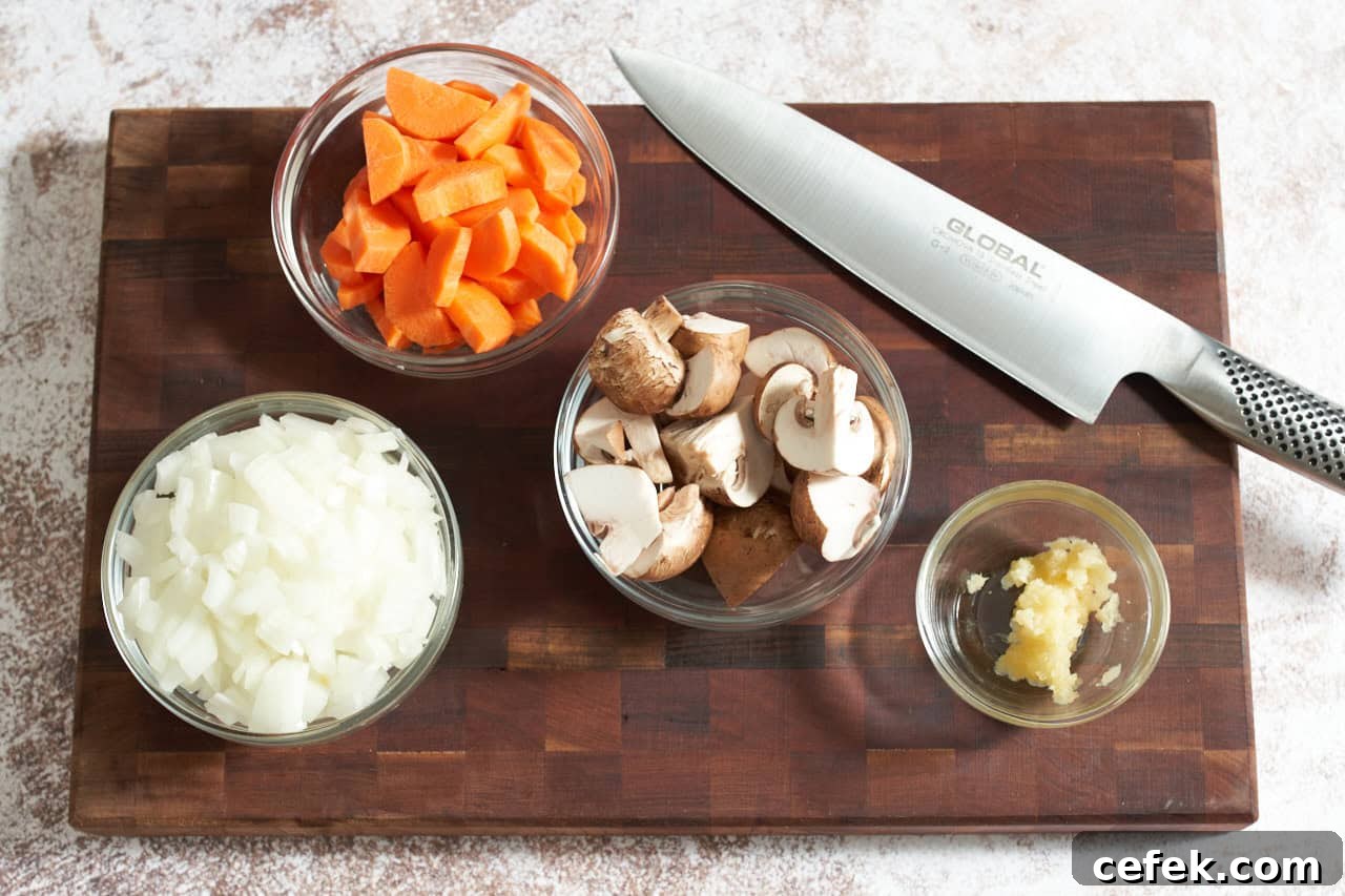 A knife on a wooden cutting board with small bowls of chopped onions, carrots, mushrooms, and garlic.