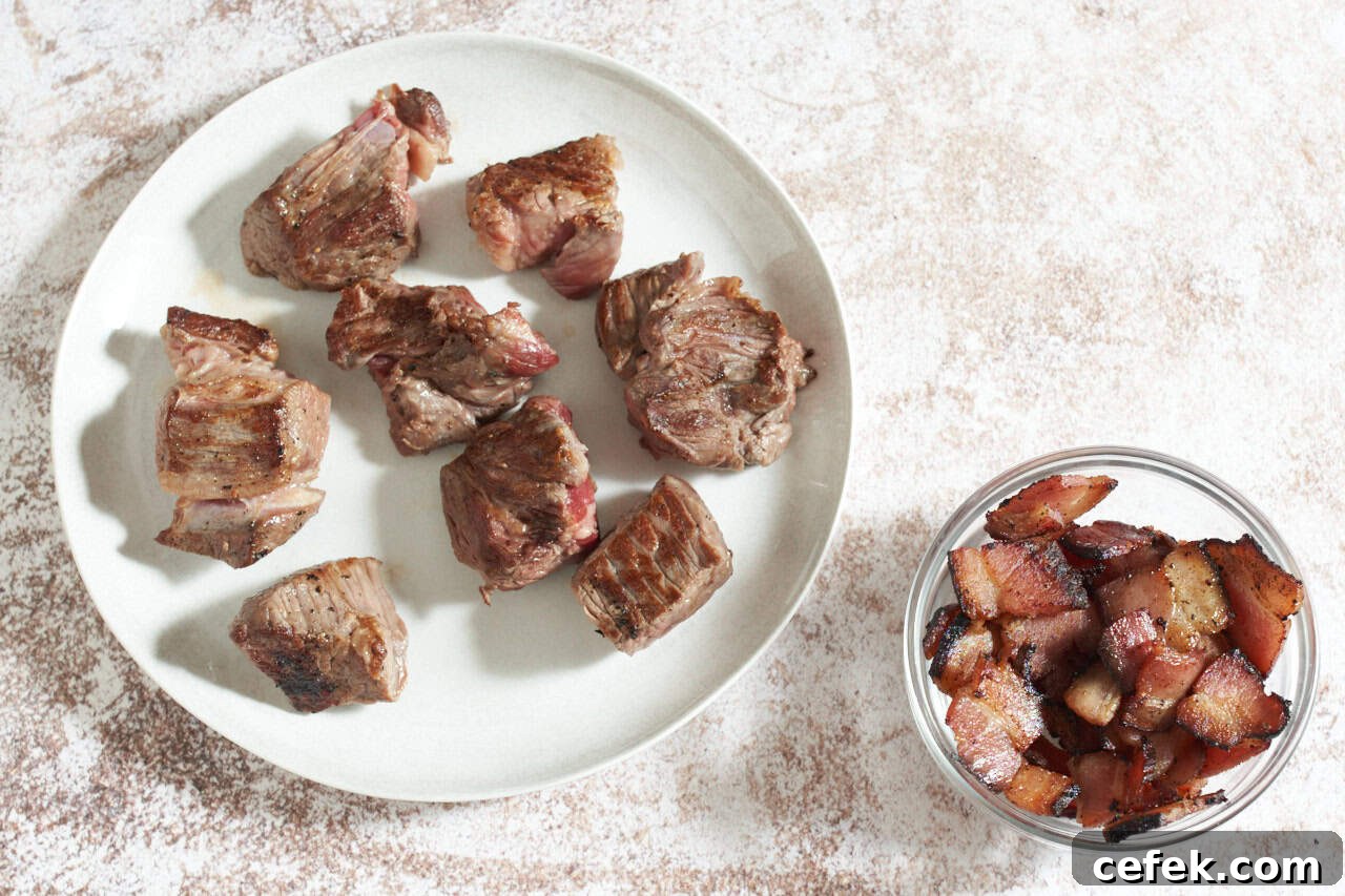 Seared beef on a white plate next to a small bowl of cooked bacon pieces.