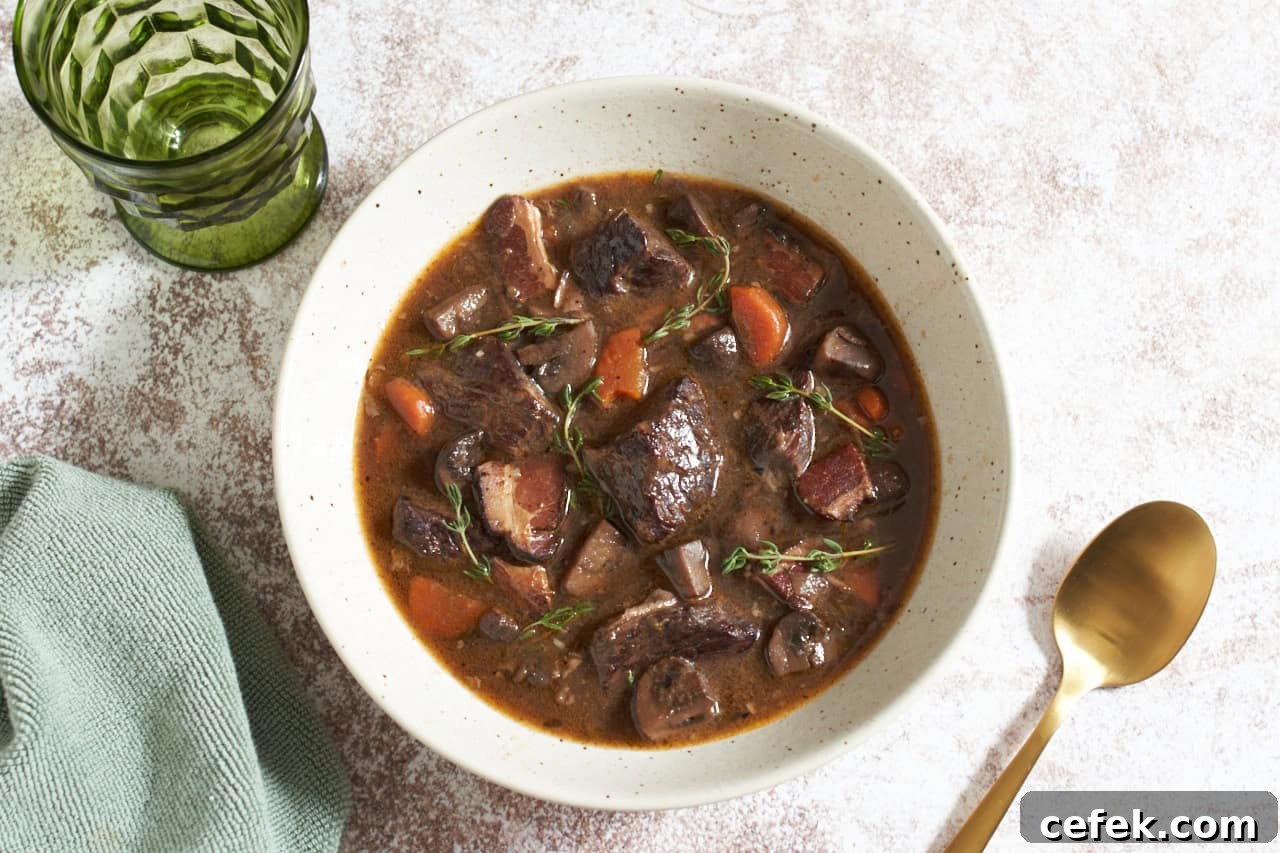 A bowl of slow cooker beef bourguignon topped with fresh thyme, a gold spoon is on the right and a green glass is in the upper left.