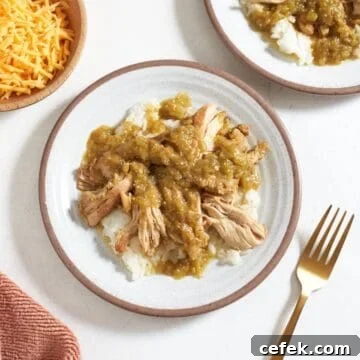 Two plates of slow cooker green chili chicken served over rice, a wooden bowl of shredded cheese is on the left, a gold fork is to the right.