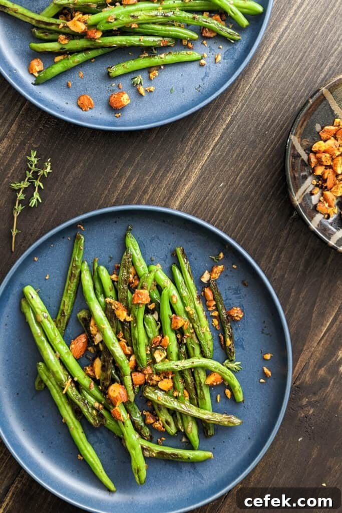 Finished serving of seared green beans topped with toasted spiced almonds on a round gray plate. The plate is on a dark brown table. At the top edge is a second finished plate and at the top edge is a bowl of toasted spiced almonds.