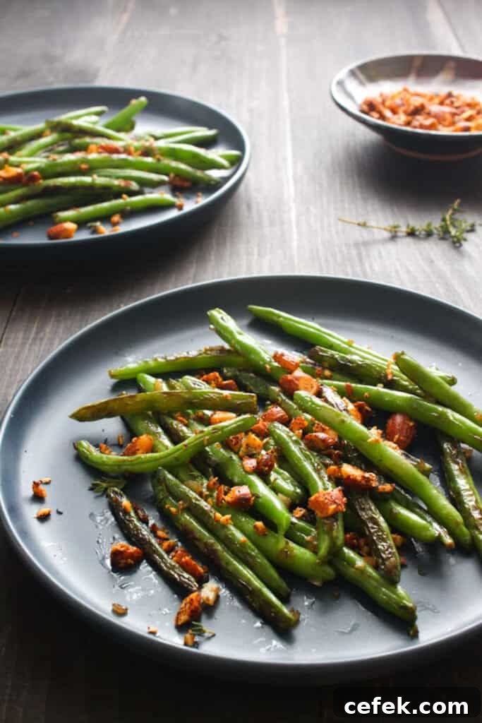 Finished serving of seared green beans with toasted spiced almonds on round gray plate. In the background is a second plate of finished green beans and a bowl of extra spiced almonds.