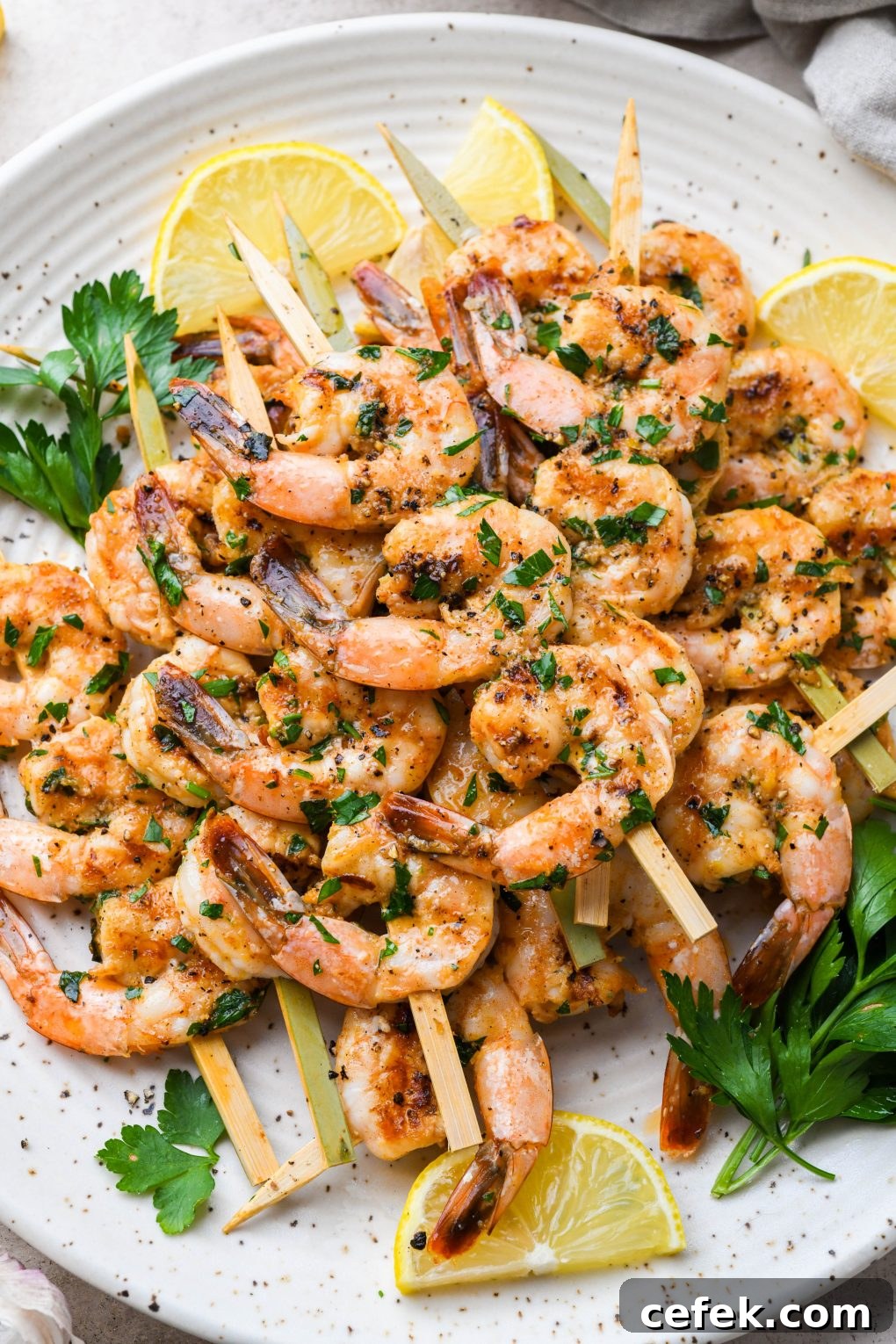 Overhead image of grilled shrimp on wooden skewers. On a light colored speckled plate topped with chopped parsley and next to some lemon wedges.