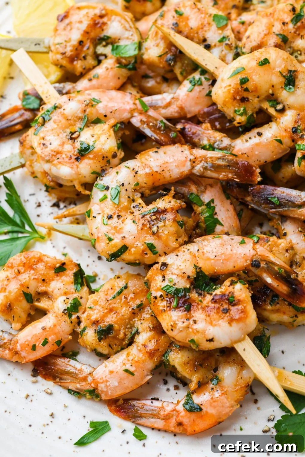 Side angle close up image of grilled shrimp on wooden skewers. On a light colored speckled plate topped with chopped parsley and next to some lemon wedges.