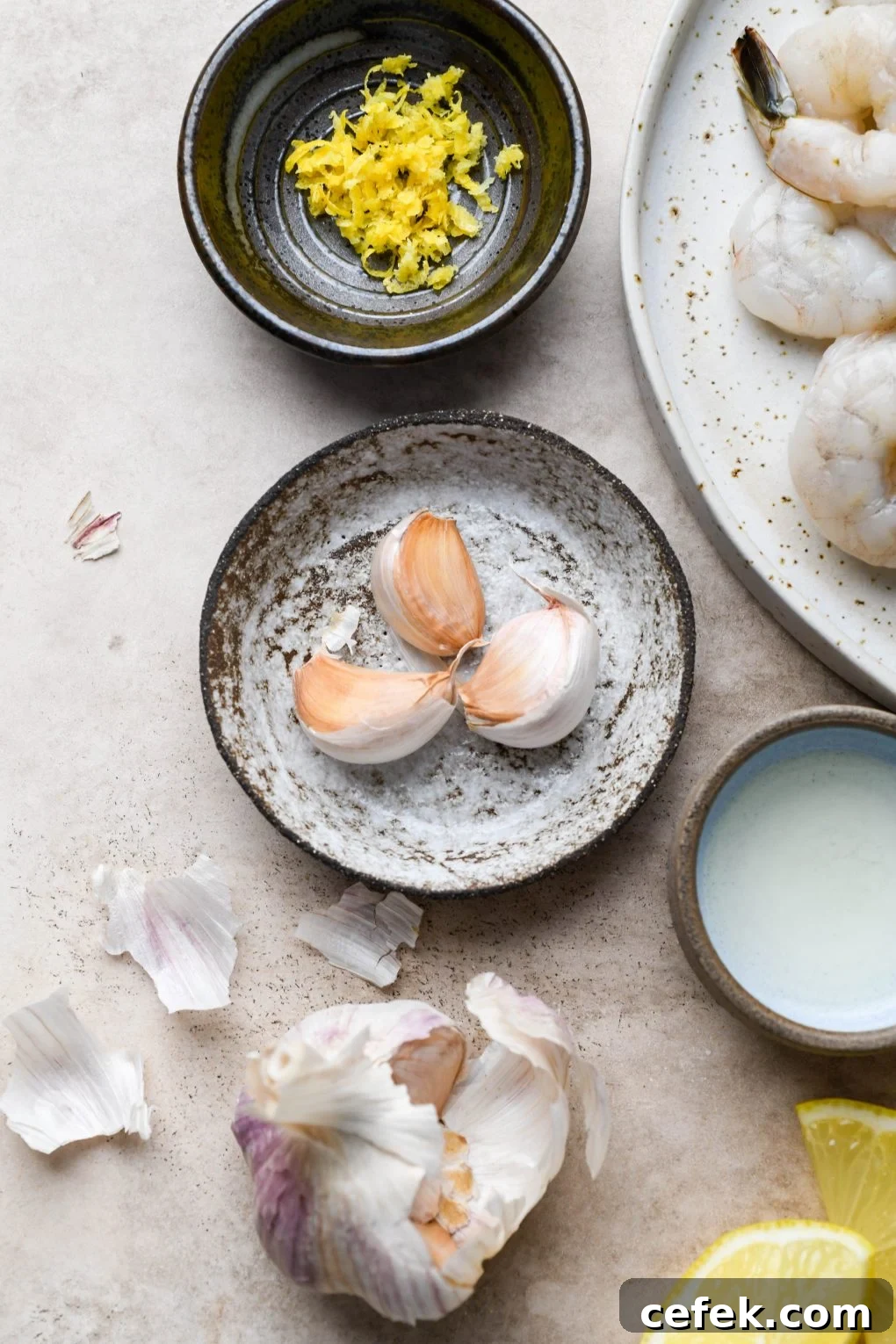 A small textured dish filled with 3 garlic cloves. Surrounded by some scattered garlic skin, a dish of lemon zest, and a small bowl of lemon juice. On a light colored background.