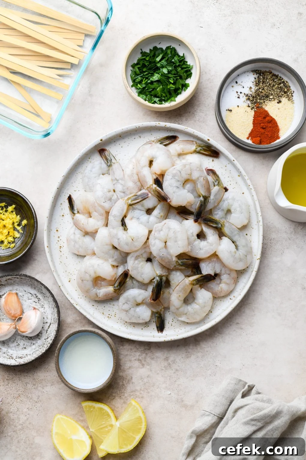 All of the ingredients for garlicky grilled shrimp skewers. Raw tail on shrimp, spices, parsley, olive oil, garlic, lemon zest, and lemon juice. On a light colored background.