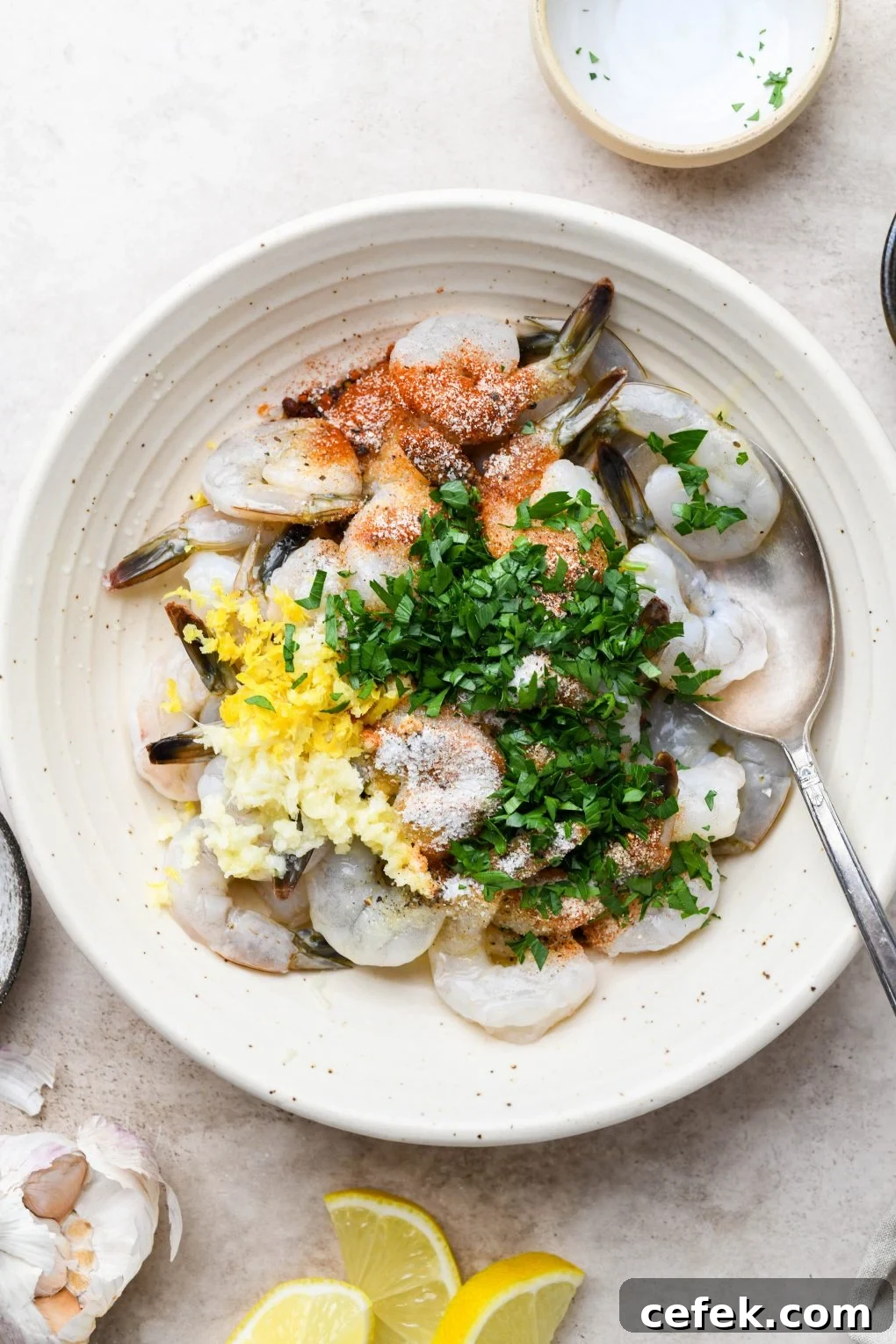 Overhead shot of a large shallow bowl with raw tail on shrimp, topped with all the ingredients for the zesty marinade - parsley, spices, lemon zest, lemon juice, and garlic.