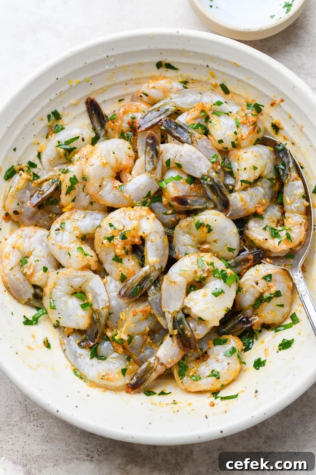 Overhead shot of a large shallow bowl with raw tail on shrimp, tossed together with all the ingredients for the zesty marinade - parsley, spices, lemon zest, lemon juice, and garlic.