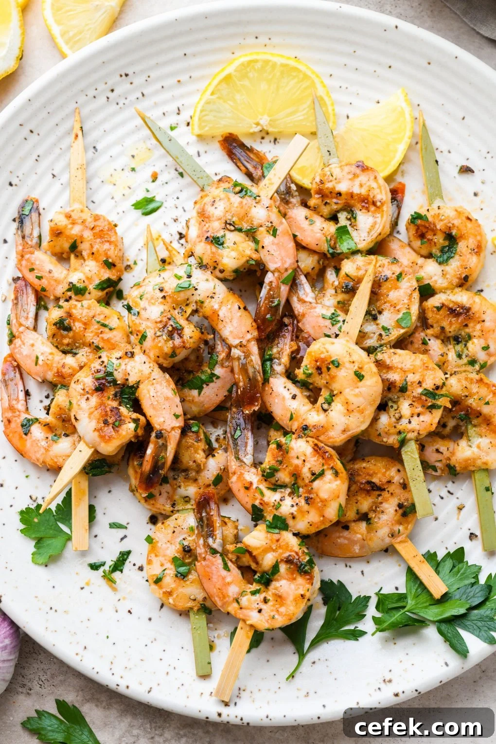 Overhead image of grilled shrimp on wooden skewers. On a light colored speckled plate topped with chopped parsley and next to some lemon wedges.