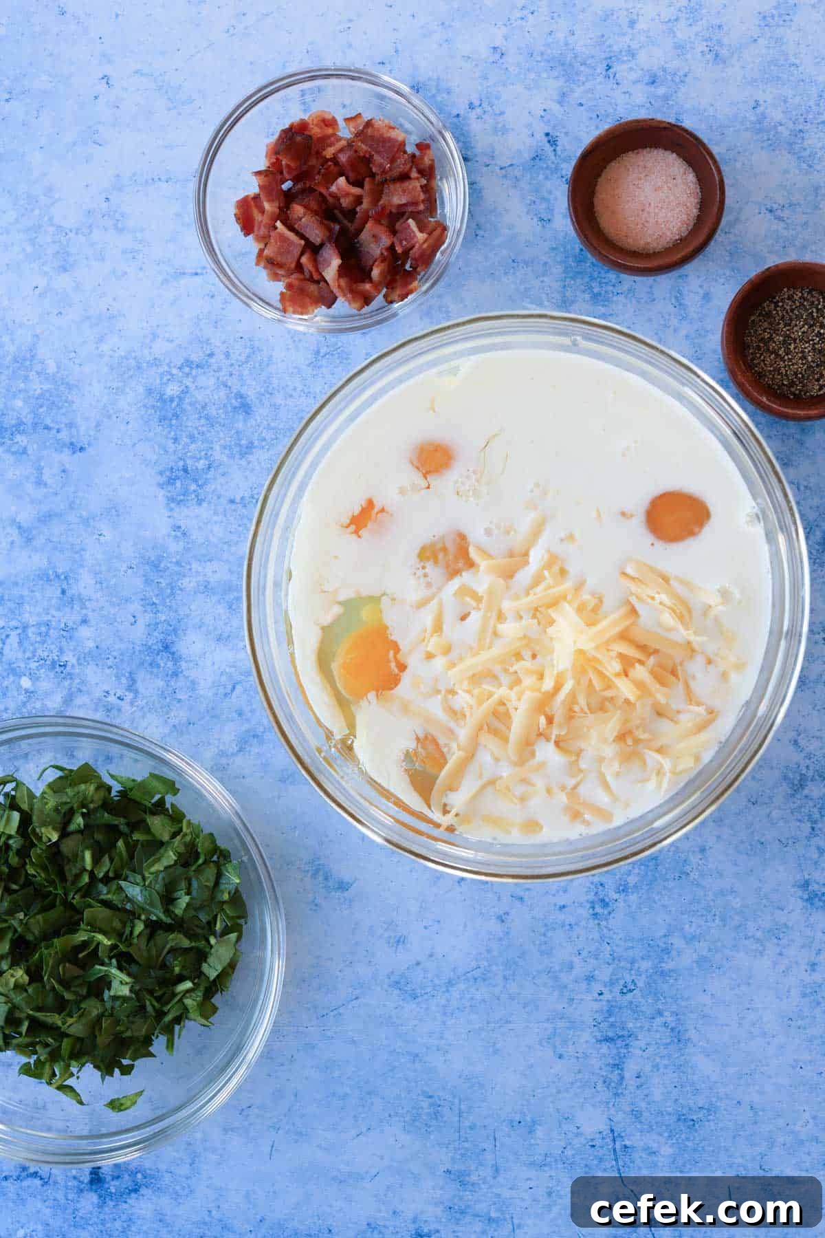 A bowl of eggs, milk, and cheese in a glass mixing bowl with other ingredients on a blue countertop.