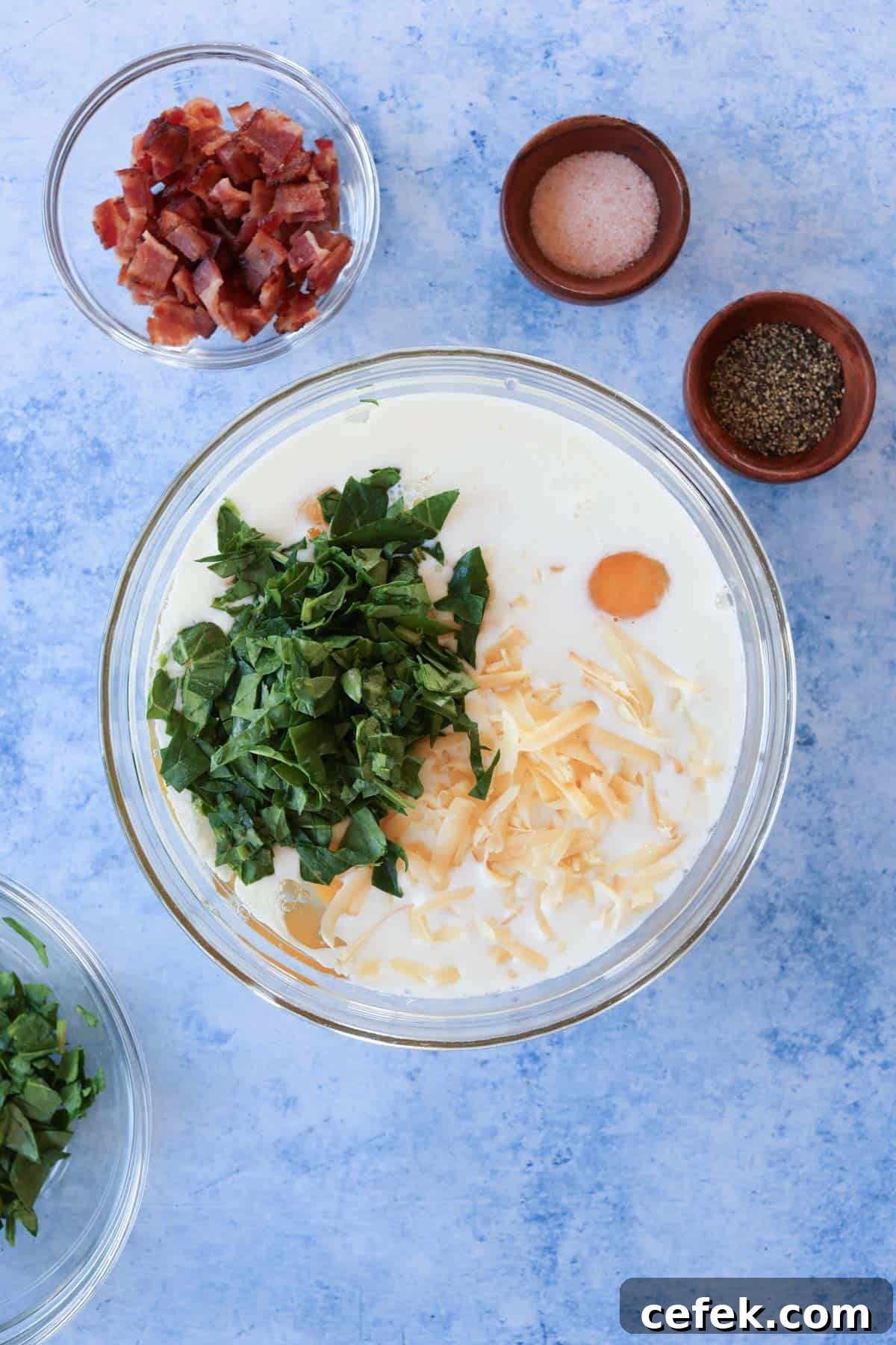 A bowl of eggs, milk, cheese and chopped spinach in a glass mixing bowl.