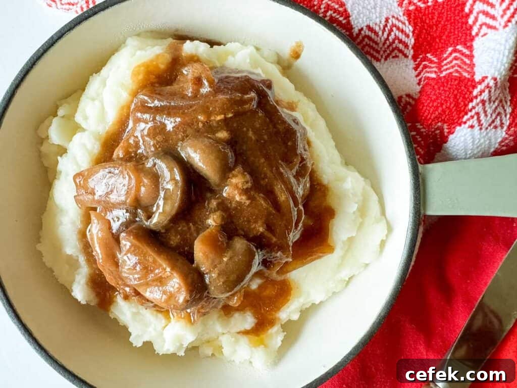 Mashed potatoes topped with brown gravy and mushrooms, reminiscent of a slow cooker Salisbury steak, in a bowl placed on a red and white checkered cloth.