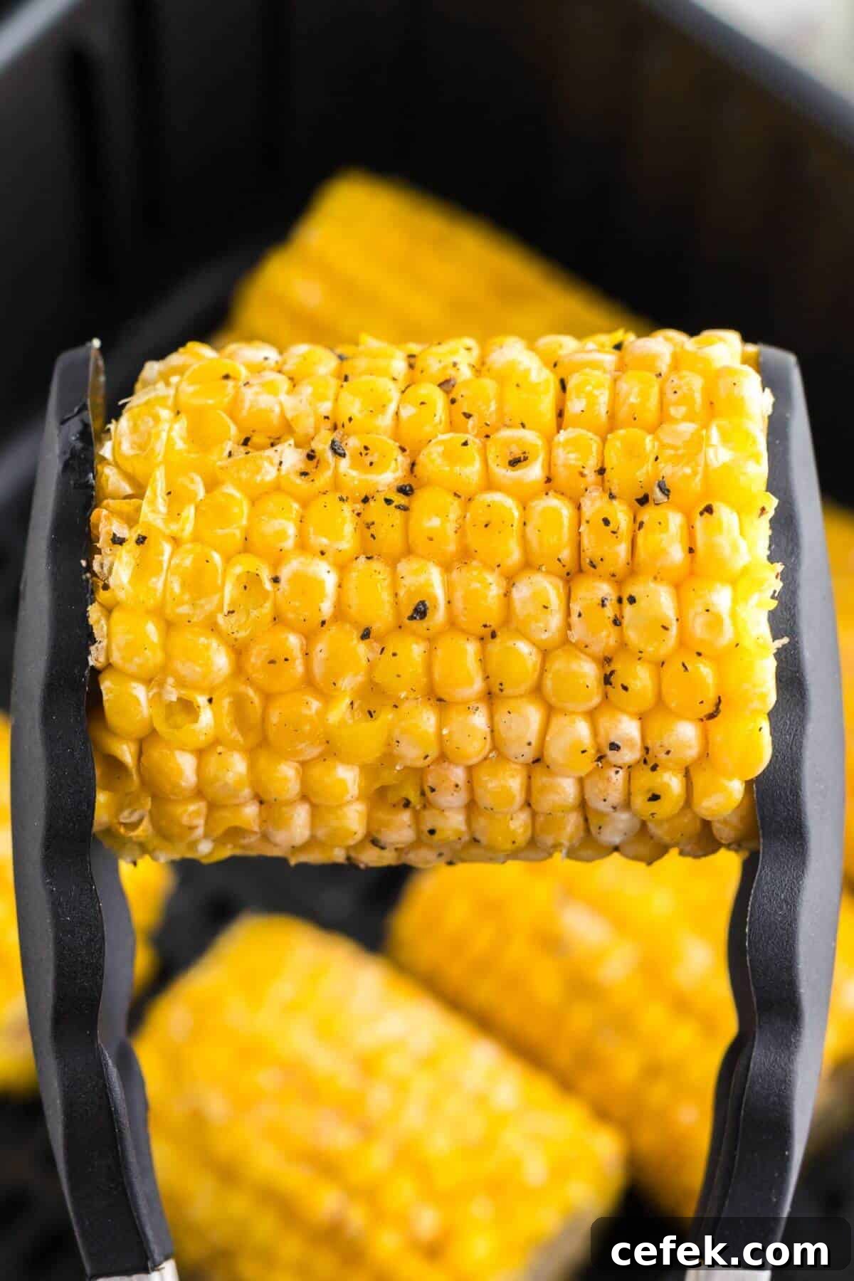 Cooked yellow corn on the cob being held with tongs and seasoned. 