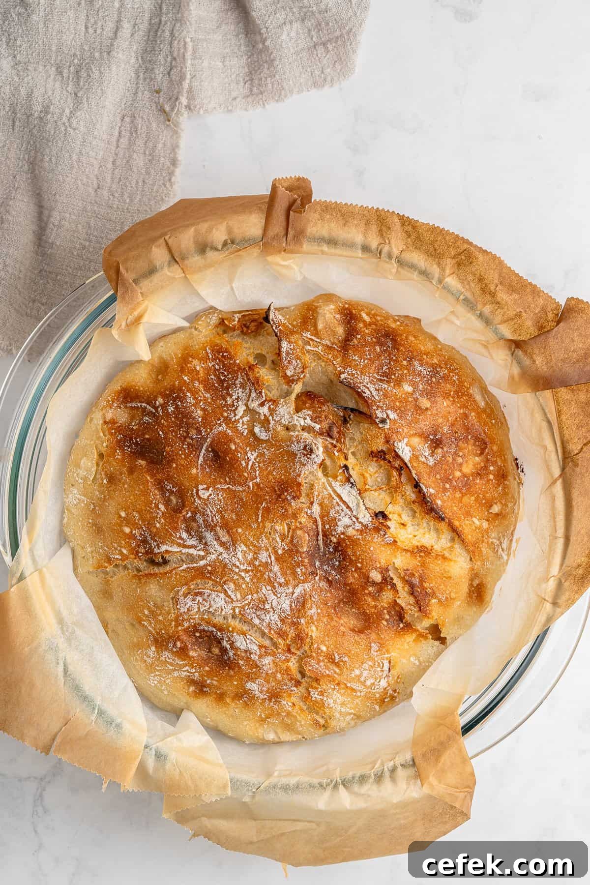 A freshly baked round loaf of crusty no knead garlic bread on parchment paper in a glass dish. The bread has a golden-brown crust with a dusting of flour. A beige cloth is partially visible in the top left corner on a light-colored surface.