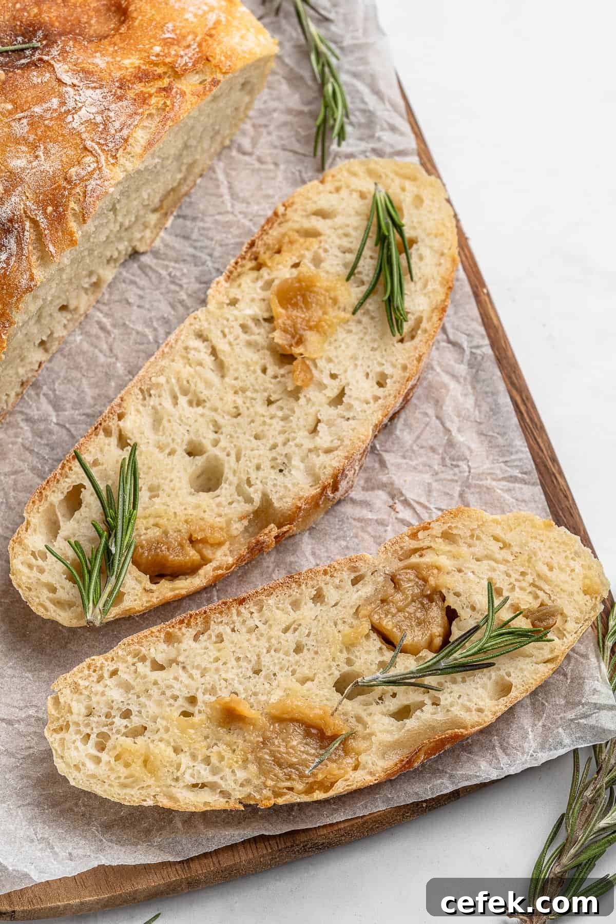 Two slices of crusty No Knead Garlic Bread topped with roasted garlic and sprigs of rosemary rest on parchment paper on a wooden cutting board. A portion of a loaf is visible in the background. Sprigs of rosemary are scattered around.