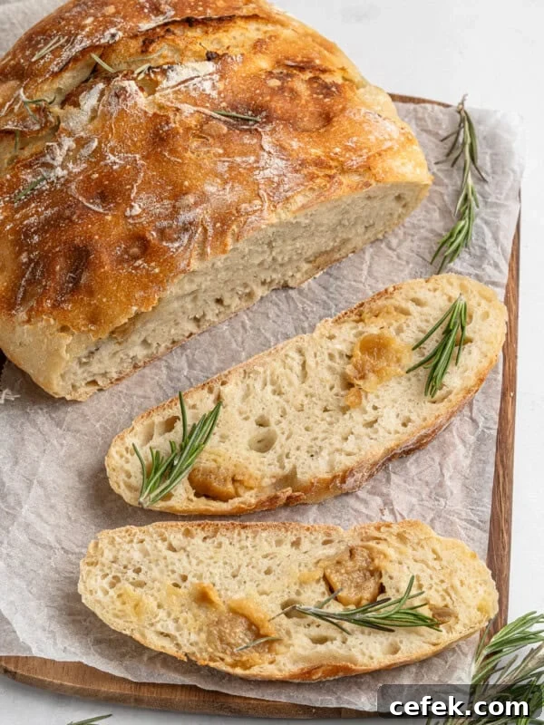Freshly baked rosemary bread on a wooden board with parchment paper. One whole round loaf and two slices are visible, garnished with sprigs of rosemary. The crust is golden and the crumb looks soft and airy.