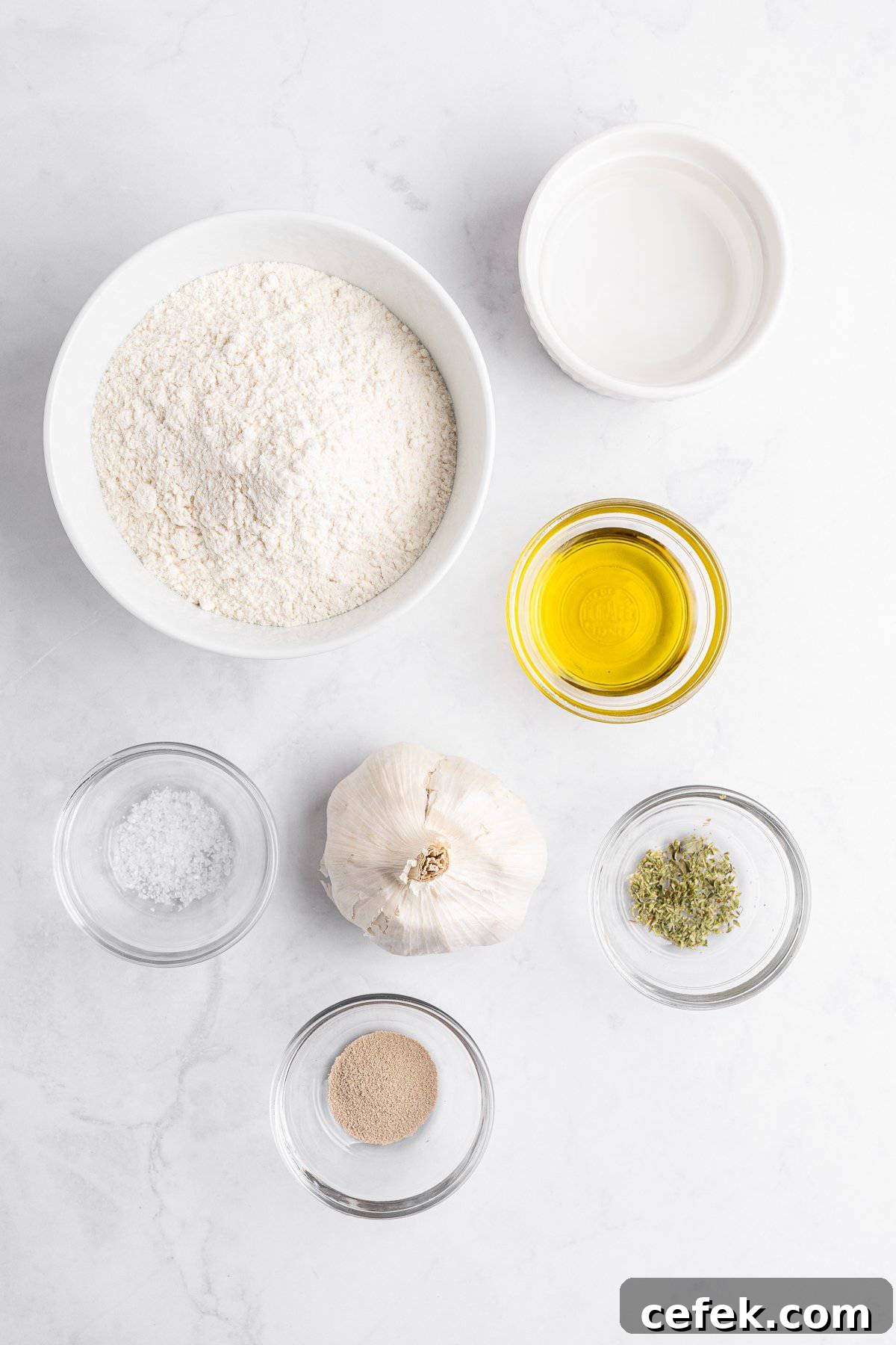 Ingredients on a white surface: a bowl of flour, a small bowl of water, a bowl of olive oil, a garlic bulb, a dish of dried herbs, a dish of salt, and a dish of yeast.