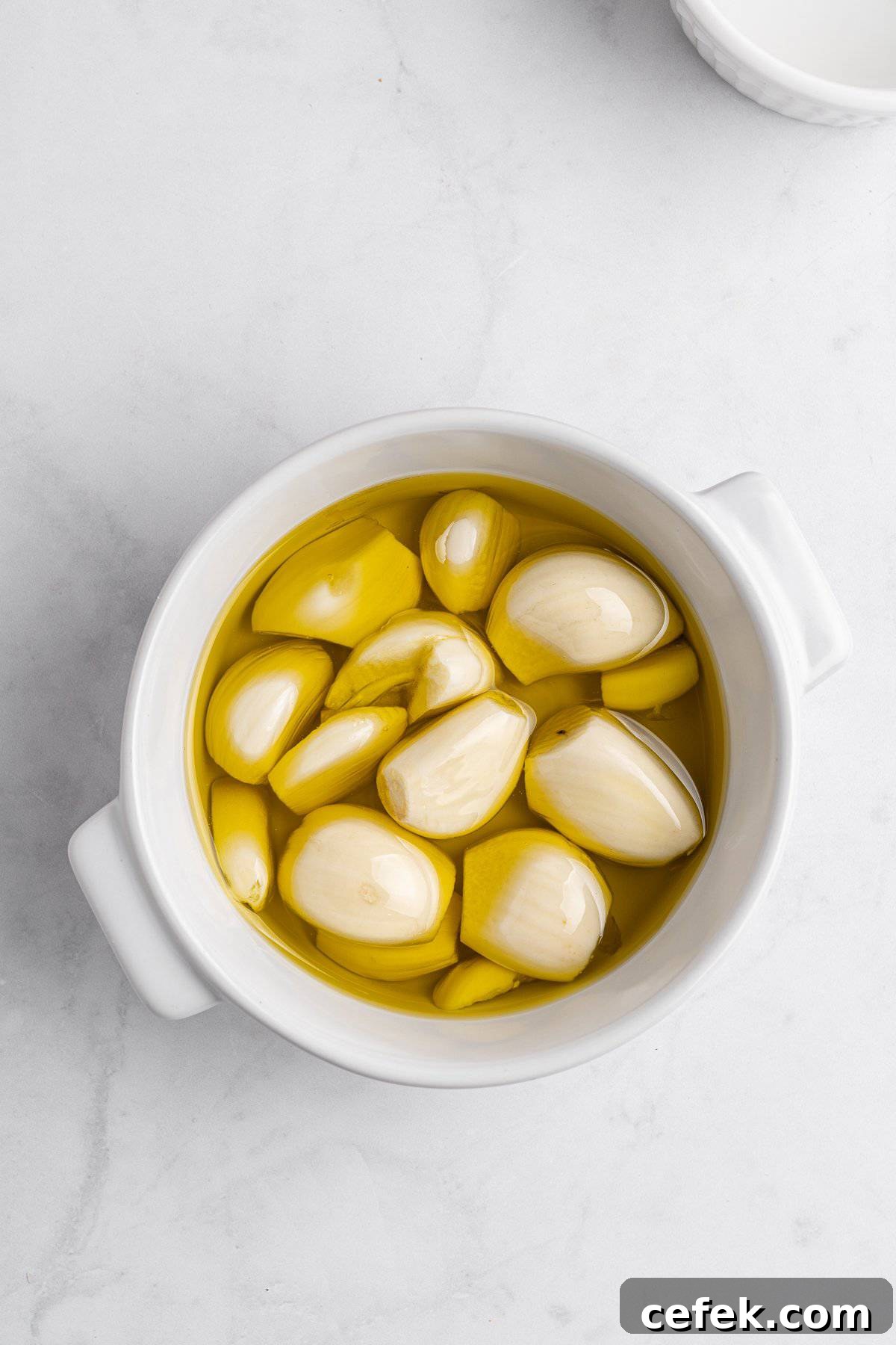 A white bowl filled with several peeled garlic cloves submerged in golden olive oil, placed on a light marble surface.