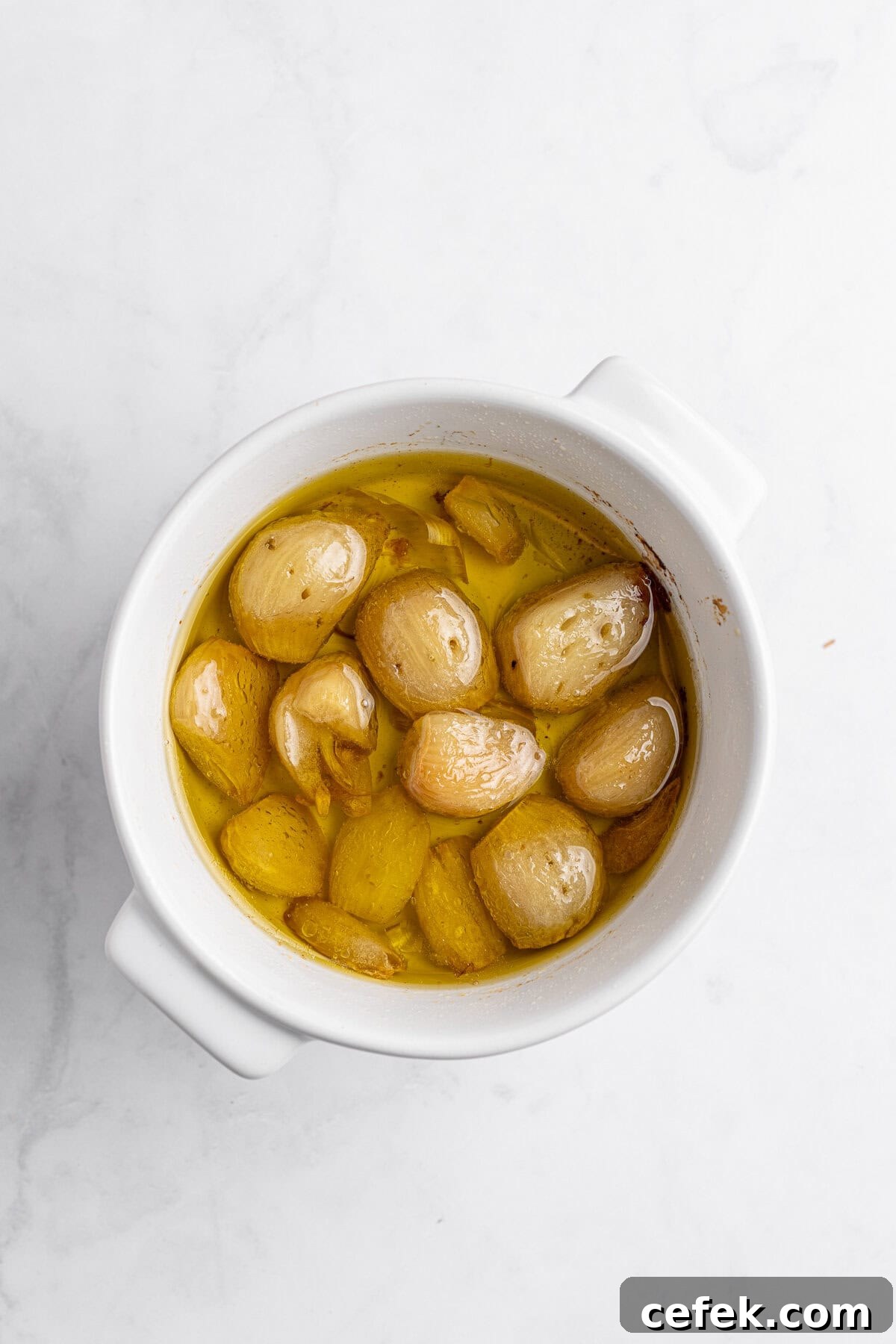 A white bowl filled with garlic cloves submerged in golden olive oil. The bowl is set against a light marble surface. The garlic appears roasted, with a soft texture and light browning.