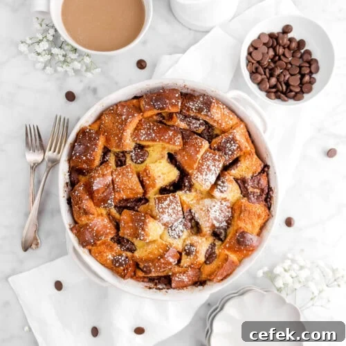 overhead shot of chocolate chip bread pudding in round casserole on white napkin with two forks, flowers, bowl of chocolate chips, cup of coffee, and white creamer beside with scalloped plates