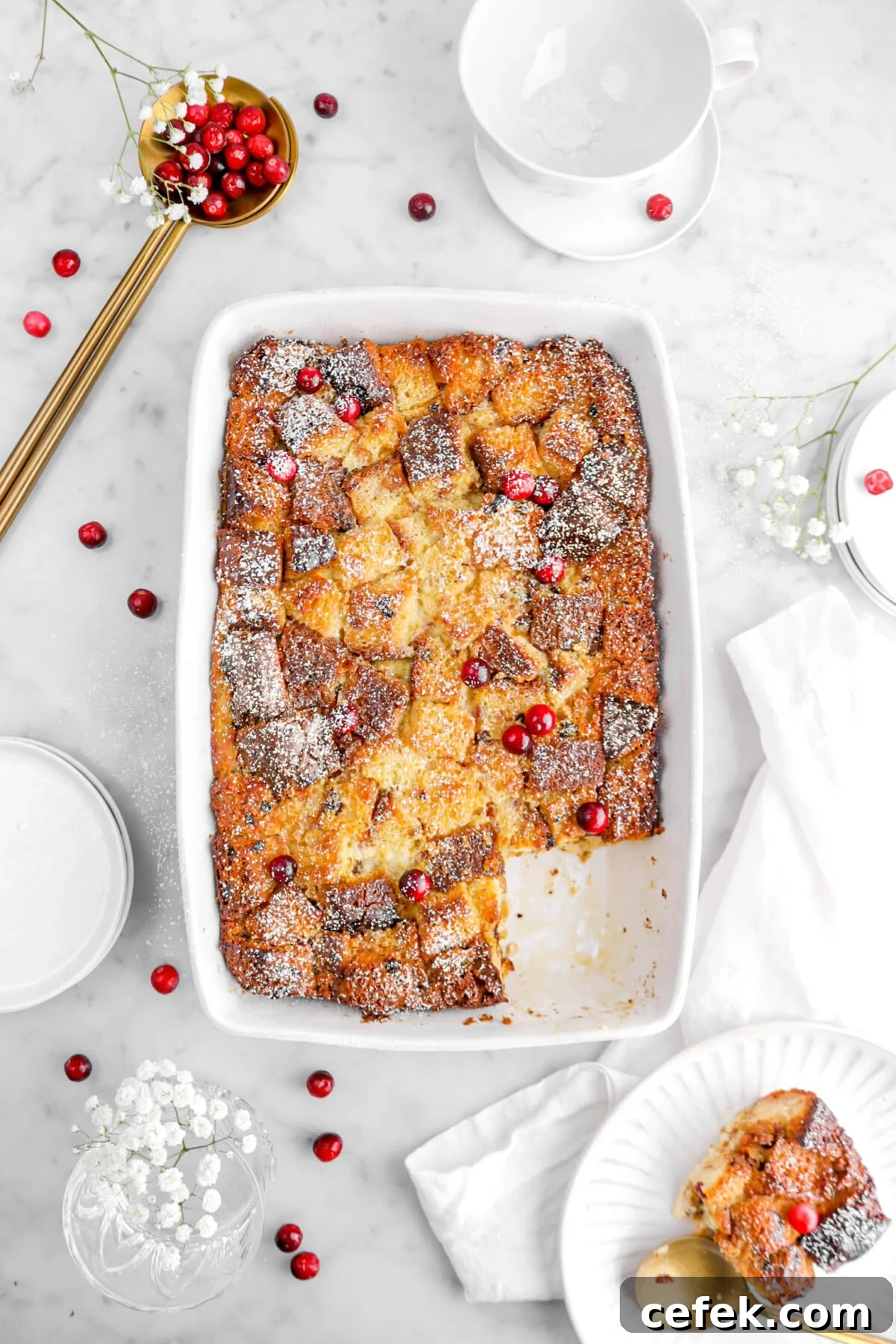 overhead shot of bread pudding with slice on white plate beside casserole with gold spoon beside, cranberries, and white flowers on marble surface.
