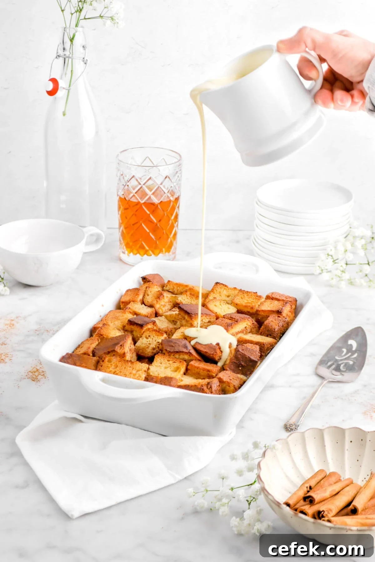 creme angalise being poured onto bread pudding on white napkim with cake server, glass of bourbon, stack of white plates, and flowers
