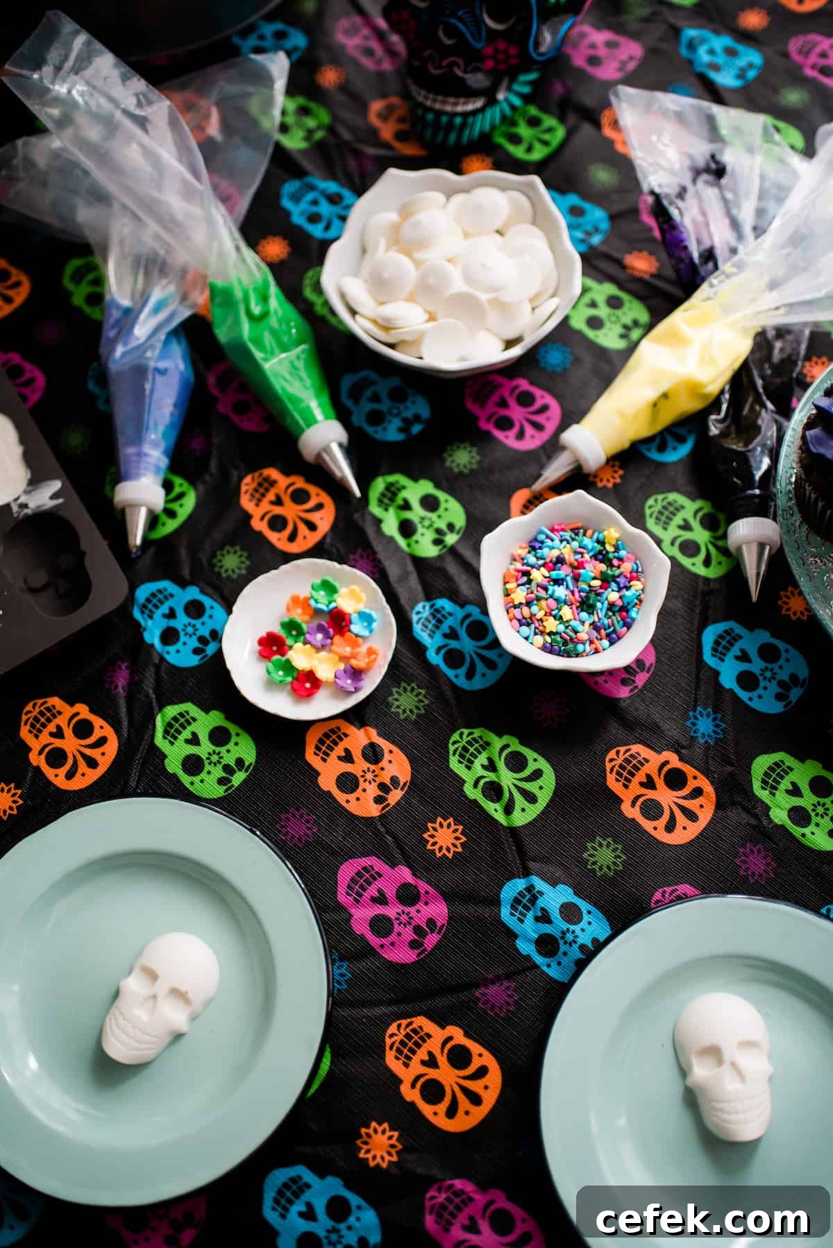 Undecorated white chocolate skulls on blue plates with piping bags filled with colorful frosting, candy flowers, and sprinkles