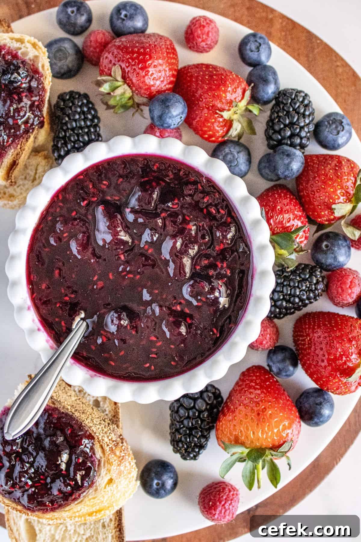 Small white dish of Mixed Berry Jam on a marble plate with some fresh berries and bread around the edge.