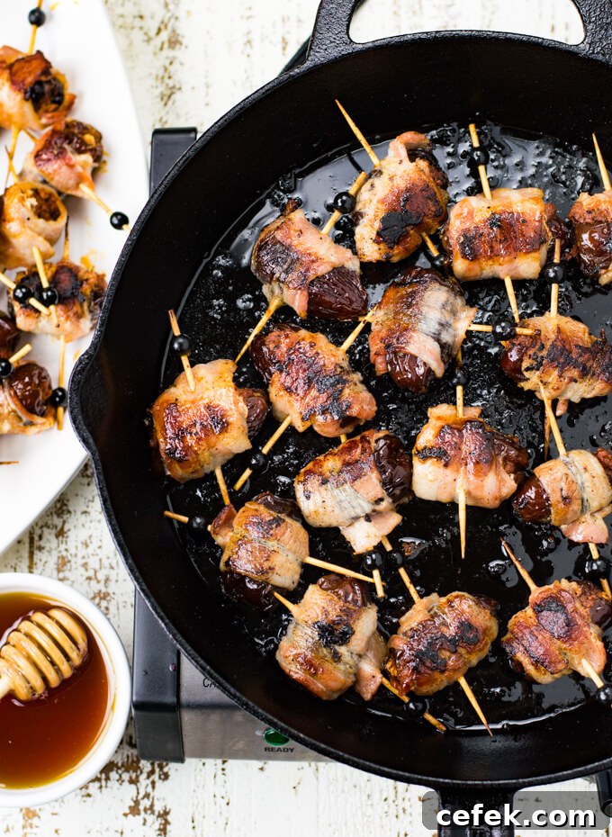 A black cast iron skillet holds cooked bacon wrapped dates stuffed with chorizo. A bowl of honey and a drizzler sits next to it.
